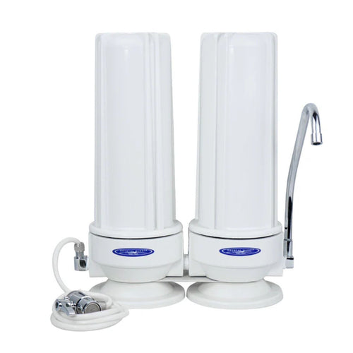 A two-cartridge water filter system with white cylindrical filter canisters, a curved chrome faucet spout, a white supply hose with metal fittings, and blue oval brand badges on the base of each canister, shown on a plain white background.