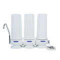 Three white water filter canisters in a row with a chrome faucet spout on the left and a white power cord on the right, shown on a plain white background.