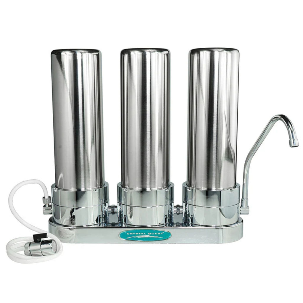 A three-cartridge stainless steel water filter pitcher with a curved metal spout, shown on a plain white background. A white plastic supply tube is attached to the left side, and a small teal label on the front reads 'STAINLESS STEEL'.
